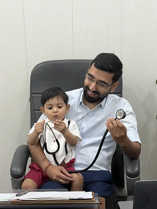 Dr. Yashasvi Verma at his clinic desk with a young child on his lap, letting him play with the stethoscope
